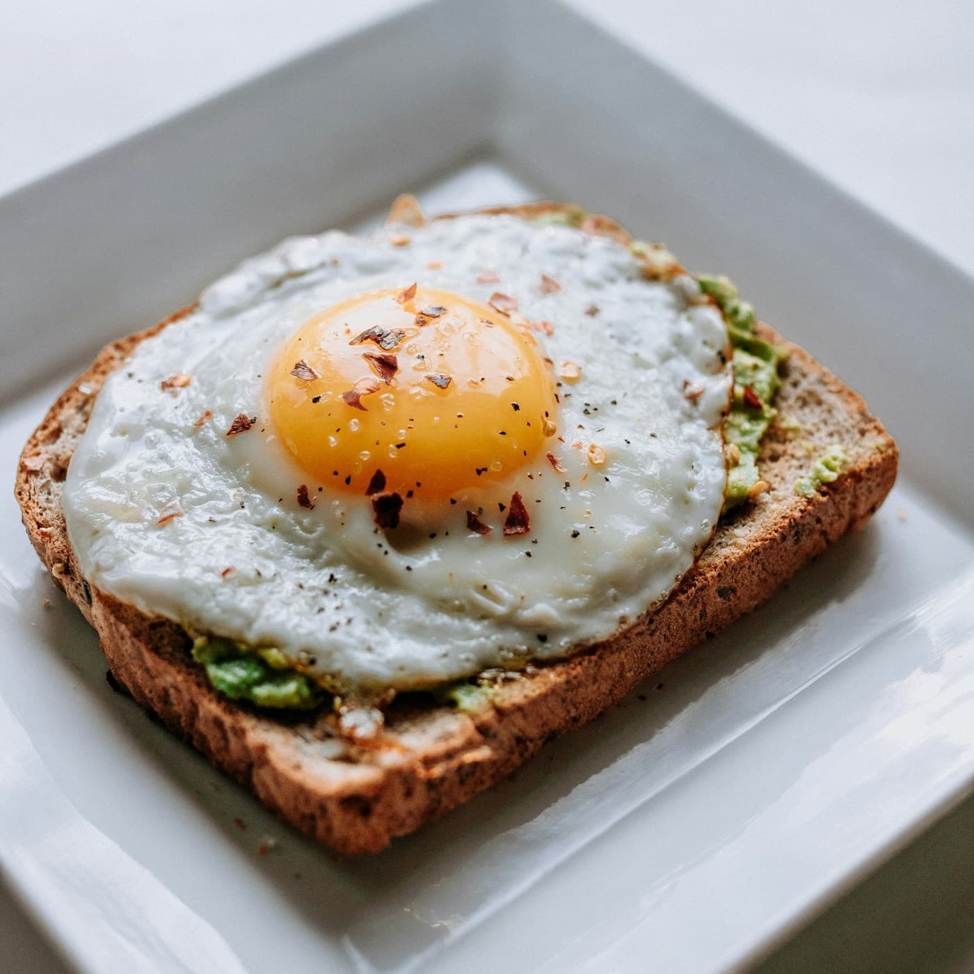 Fried egg sprinkled with chilli flakes on avocado-covered toast on a white plate