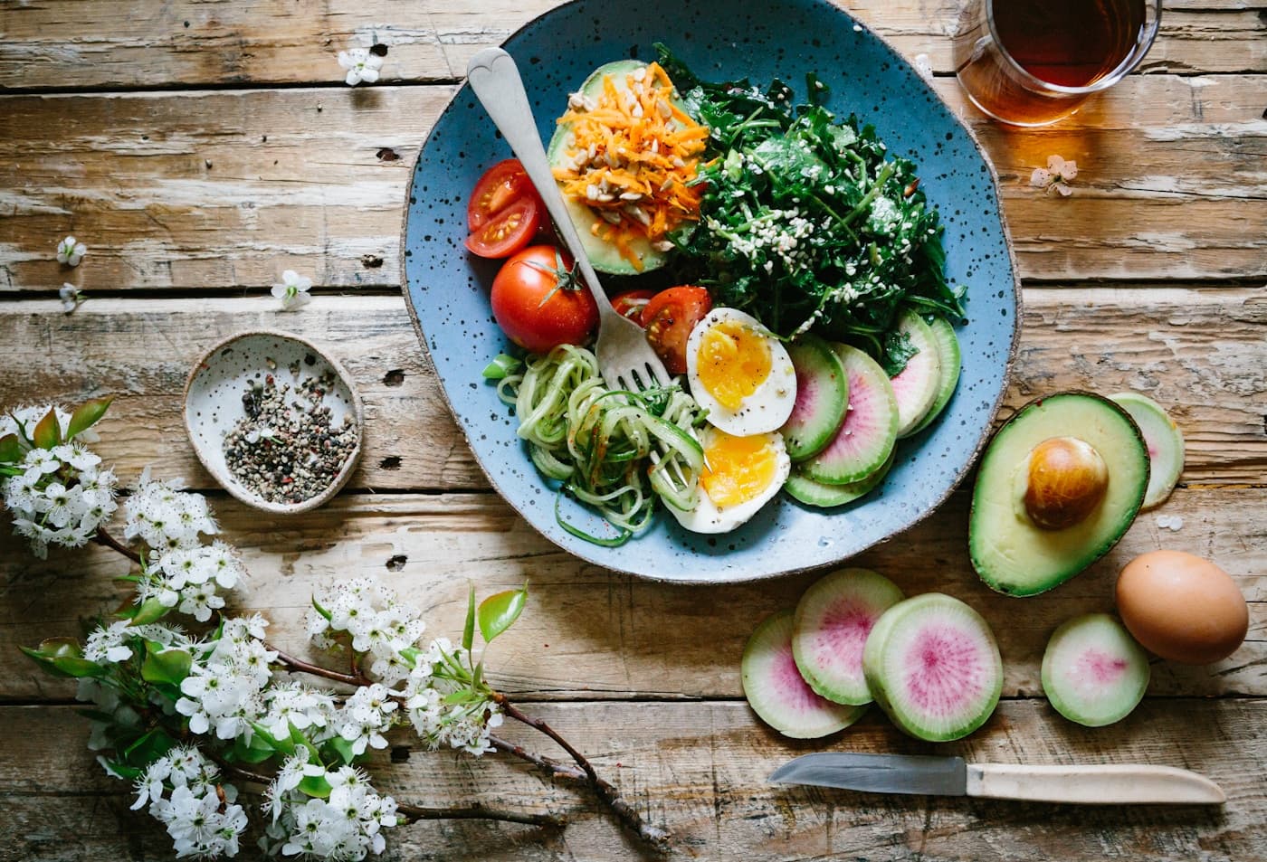 A blue bowl with halved eggs, greens, avocado, tomatoes, cucumber ribbons, and sliced radish